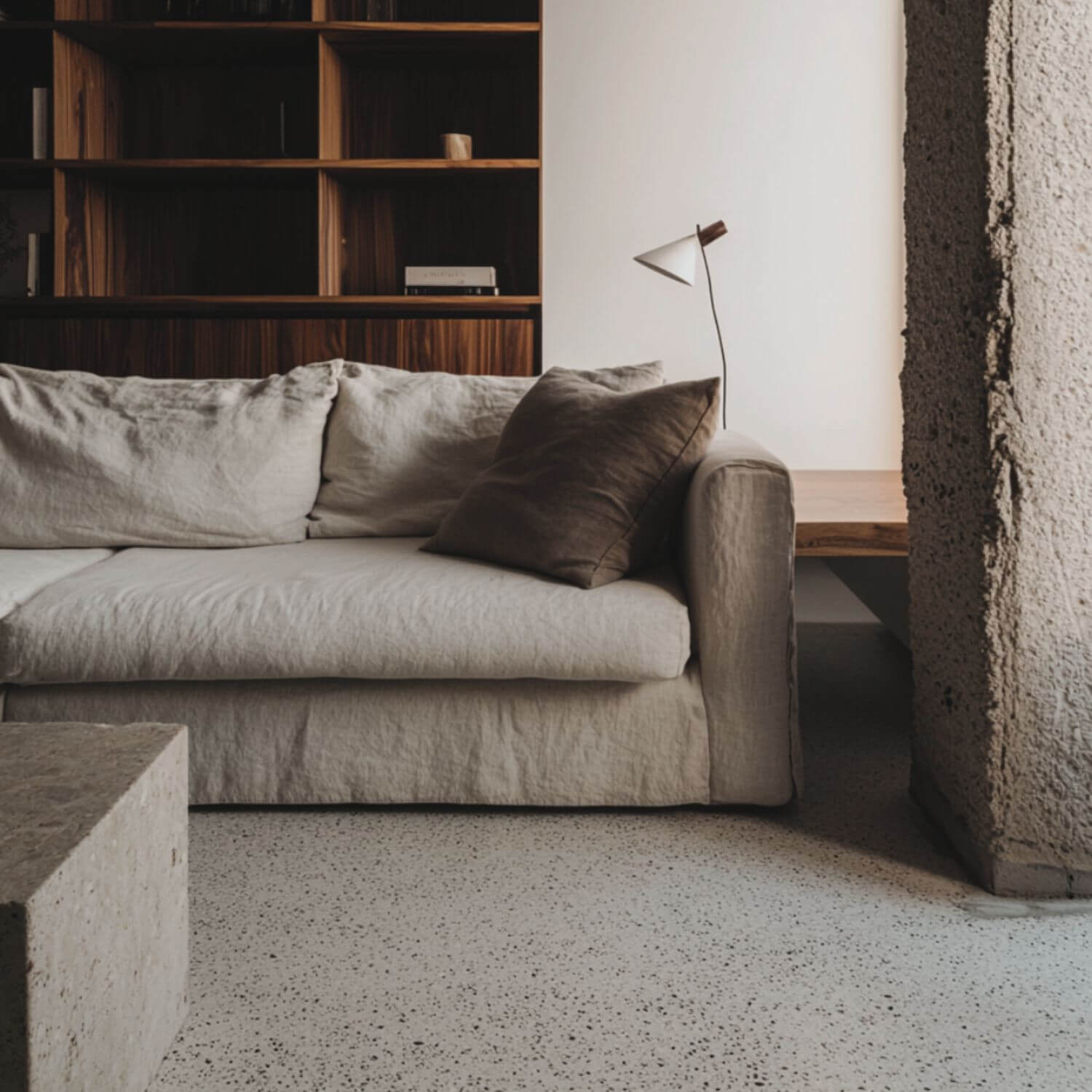 Salón reformado con suelo de terrazo original, sofá en lino beige arrugado, cojín oscuro y estantería de madera al fondo.